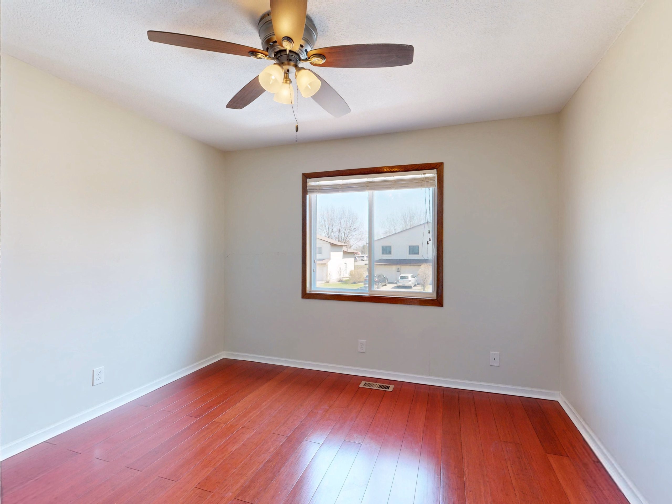 Spacious empty room with wooden flooring, window view, and ceiling fan.